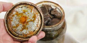 Hand holds rusty tin lid with black stains. Opened glass jar with homemade pickled Suillus.